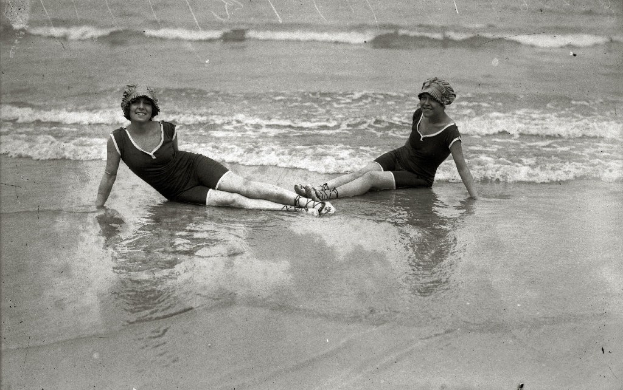 Zwei Frauen in Badesachen und Hüten sitzen lachend im flachen Wasser am Strand, genießen die Sonne und die Wellen in einem zeitlosen Schwarz-Weiß-Bild.