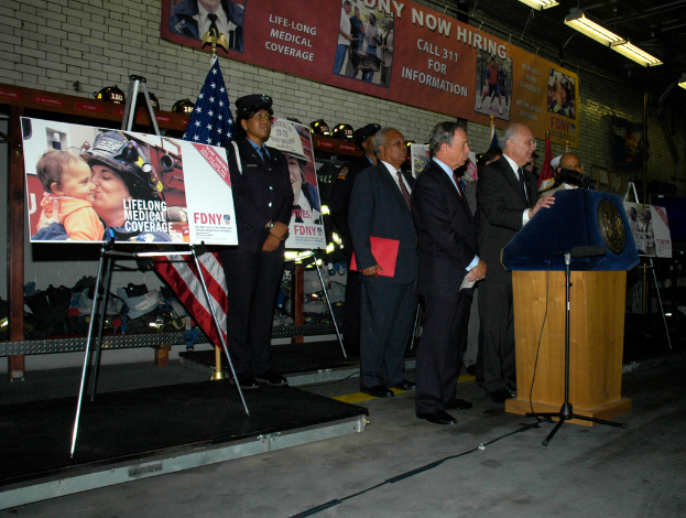 Eine Gruppe von Menschen steht in der Nähe eines Podiums mit Mikrofonen, umgeben von Tafeln auf Ständern, Flaggen und Wandtafeln, alles unter hellen Scheinwerfern.