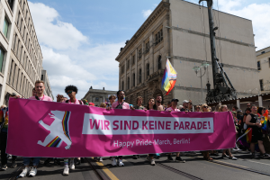 Eine Gruppe von Menschen marschiert auf einer Straße in Berlin, Deutschland, mit einer pinken "Happy Pride March"-Fahne, mit Gebäuden auf beiden Seiten und einem Fahnenmast im Vordergrund unter einem bewölkten Himmel.