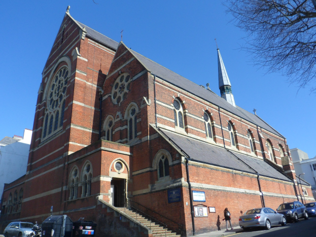 Eine große Backsteinkirche mit einem Kirchturm, umgeben von Gebäuden, Straßenmasten, Straßenlaternen, Kraftfahrzeugen, Fußgängern, Mülltonnen, Bäumen und einem klaren blauen Himmel.
