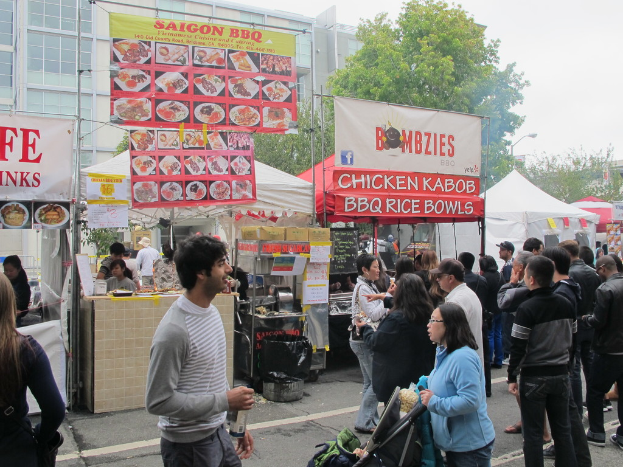 Menschen stehen auf einer Straße vor der Stadt, ein Mann in einem grauen und weißen T-Shirt hält eine Wasserflasche und eine Frau in einer blauen Jacke hält einen Kinderwagen. Hinter ihnen sehen wir Stände mit weißen und roten Zelten, farbige Banner und Gebäude, Bäume und eine Straßenlaterne im Hintergrund.