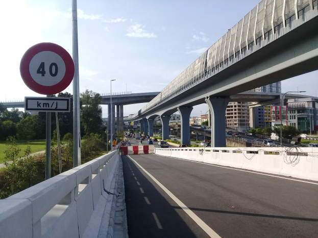Autobahn mit Tempolimit-Schild, Fahrzeugen, Brücke mit Pfeilern, Laternen, Bäumen, Gebäuden und bewölktem Himmel im Hintergrund.
