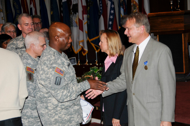 Eine Gruppe von Menschen, die mit zwei Personen Handschlag geben, Fahnen im Hintergrund, ein Podium mit Mikrofon und eine Topfpflanze in der Nähe.