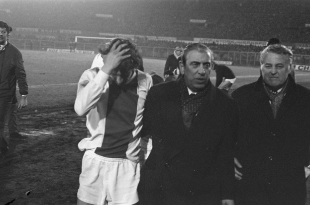 Eine Gruppe verzweifelter Männer in Schwarz-Weiß-Kleidung steht zusammen auf einem Fußballfeld, im Hintergrund ein Stadion.