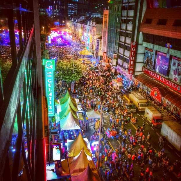 Eine bunte KarnevalsSzene mit einer großen Menge auf der Straße, umgeben von Zelten, Geschäften, Gebäuden und Bannern, mit zahlreichen Lichtern im Hintergrund.