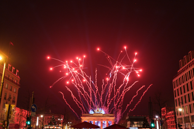 Eine belebte Stadtstraße an einem Silvesterabend in Berlin mit Gebäuden, Bäumen, Laternenmasten, Verkehrszeichen, Zeltplanen, Menschen und einem beeindruckenden Feuerwerk, das den Himmel erhellt.