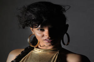 Eine Frau in einem goldenen Kleid und großen Kreolen steht selbstbewusst vor einer Wand, ihr glatter Bob und natürliches Make-up unterstreichen ihren entschlossenen Gesichtsausdruck, während sie direkt in die Kamera schaut.