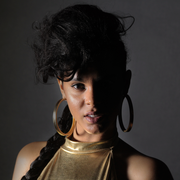 Eine Frau in einem goldenen Kleid und großen Kreolen steht selbstbewusst vor einer Wand, ihr glatter Bob und natürliches Make-up unterstreichen ihren entschlossenen Gesichtsausdruck, während sie direkt in die Kamera schaut.