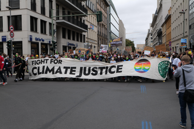 Eine Gruppe von Menschen marschiert mit einem 'Kampf für Klimagerechtigkeit'-Schild die Straße entlang, mit Gebäuden, Schildern, Ampeln und Bäumen an der Straße unter einem bewölkten Himmel.
