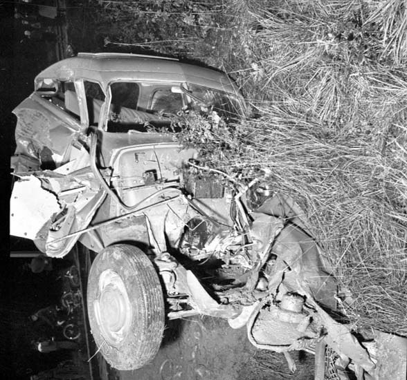 Schwarzes und weißes Foto eines schwer beschädigten Autos mit eingedellter Front und zersplitterten Fenstern am Straßenrand, mit einem Baum und verstreuten Gegenständen im Hintergrund.
