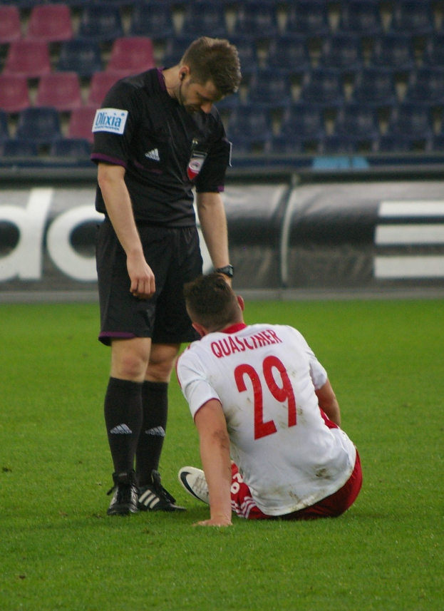 Ein Fussballspieler in Sportkleidung sitzt neben einem Schiedsrichter in einer Stadionumgebung mit Plakaten und Stühlen im Hintergrund.