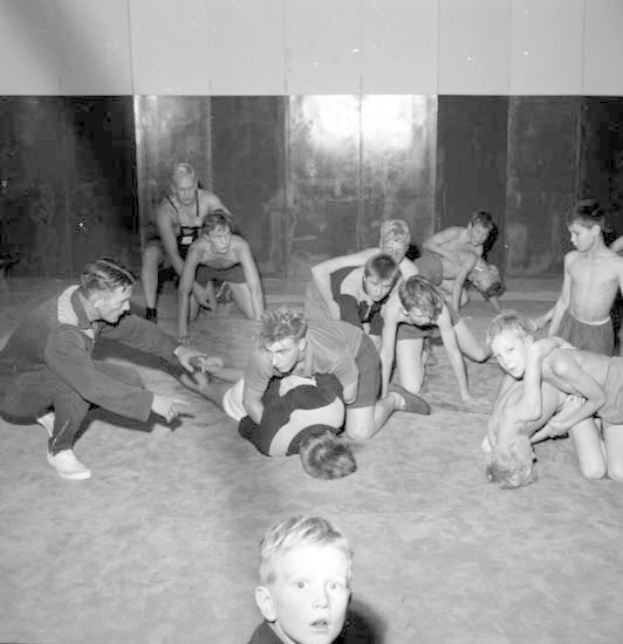 Schwarz-weißes Foto von jungen Jungs, die in einer Turnhalle miteinander ringen, mit einer Wand im Hintergrund.