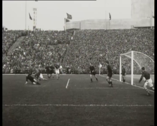 Ein Schwarz-Weiß-Foto eines WM-Finalspiels 1958 zwischen Manchester United und Liverpool mit Spielern auf dem Feld, einem Torpfosten auf der rechten Seite, Zuschauern auf den Rängen und Fahnen im Hintergrund.