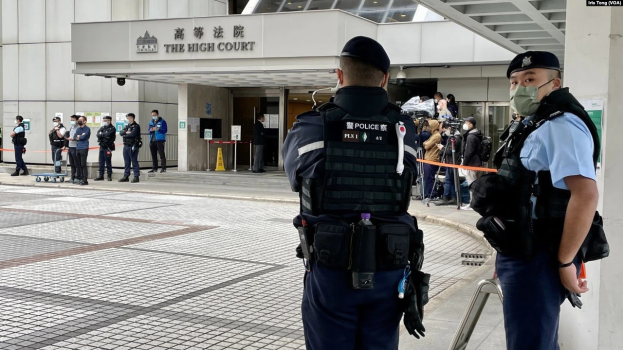 Zwei Polizisten in Uniform, Mützen und Masken stehen vor einem Gebäude am Hong Kong International Airport, umgeben von einer Gruppe von Menschen, einige mit Kameras, mit Pfosten im Hintergrund, die mit Bändern geschmückt sind.