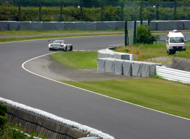 Ein Rennwagen fährt eine Rennstrecke entlang, im Hintergrund ein Polizeiauto, umgeben von Grünfläche und Absperrungen, mit Text unten.