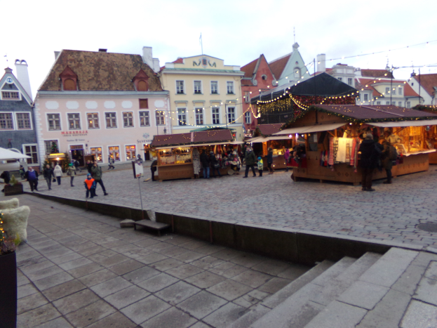 Ein belebter Weihnachtsmarkt in Tallinn, Estland mit Menschen um geschmückte Stände, festlich geschmückte Gebäude im Hintergrund, bewölkter Himmel, Treppe zum Markt und Topfpflanzen.