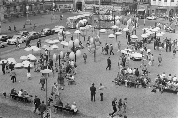 Ein Schwarz-Weiß-Foto eines belebten Stadtplatzes mit Menschen, die gehen, auf Bänken sitzen und Fahrzeugen sowie Gebäuden mit Fenstern und Namensschildern im Hintergrund.