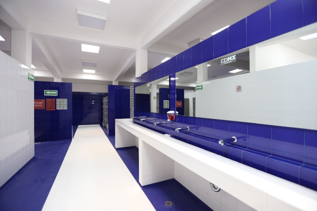 Öffentliche Toilette mit blauen Kachelwänden, weißen Tresen, Waschbecken mit Wasserhähnen, einer Plattform mit einer Flasche und anderen Gegenständen, Texttafeln an den Wänden, Deckenleuchten und Säulen.