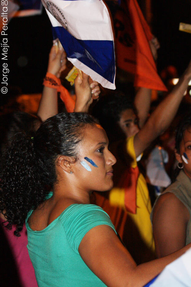 Eine Frau in einem grünen T-Shirt steht bei einer Demonstration und hält ein weißes Schild, während viele andere Menschen hinter ihr stehen.