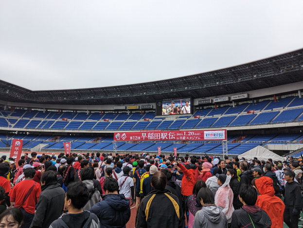 Eine große Menschenmenge steht vor einem Stadion während der Eröffnungszeremonie der Olympischen Spiele 2020 in Tokyo. Im Hintergrund sind Banner, Stühle und ein Bildschirm zu sehen. Der Himmel ist oben sichtbar und es gibt ein Wasserzeichen in der rechten unteren Ecke.