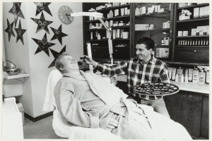 Ein Mann lächelt in einem Zahnarztstuhl, während ein anderer Mann neben ihm steht und einen Teller hält, mit einem Waschbecken, einer Uhr an der Wand und Regalen mit Flaschen im Hintergrund.