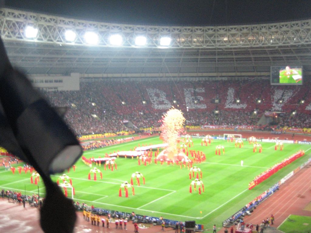 Ein Stadion mit Flutlicht, Eisengittern, Zuschauern, stehenden Individuen, Werbetafeln, Anzeigetafeln, Sicherheitskameras, Flaggen und Dekorationen.