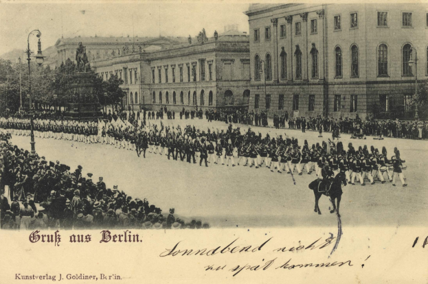 Ein Schwarz-Weiß-Foto einer Parade in Berlin, Deutschland, mit Menschen auf dem Boden und auf Pferden, einer Statue, Straßenlaternen, Bäumen, Gebäuden und einem bewölkten Himmel, mit Text am unteren Rand.