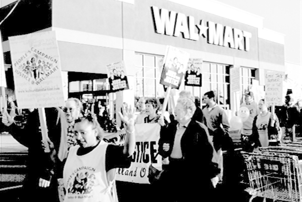 Ein Schwarz-Weiß- Bild von Menschen, die protestieren und Plakate halten, mit einem Einkaufszentrum im Hintergrund.