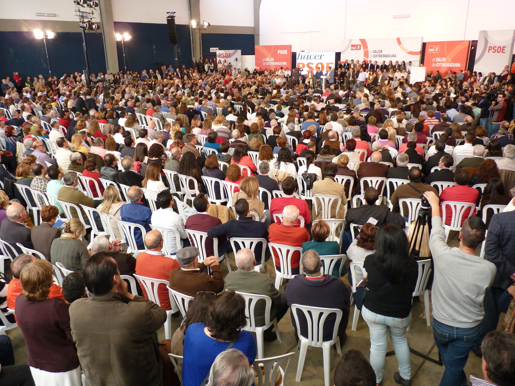 Ein volles Stadion mit vielen sitzenden und stehenden Menschen, eine Bühne mit zwei Rednern am Podium und mehreren Fahnen und Lichtern in der Nähe.