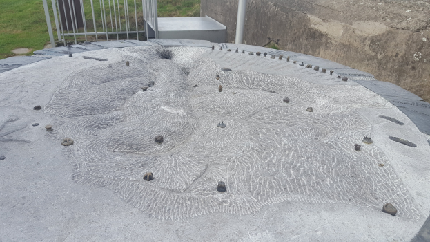 Große Betonstruktur mit einem kreisförmigen Design oben drauf, umgeben von einem Metallzaun, einem Pfahl, einer Wand und Gras.