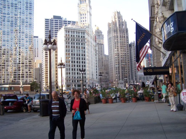 Eine belebte Straßenszene mit Menschen, Blumentöpfen, Autos, Straßenlaternen, Gebäuden, Geschäften und Fahnen auf der rechten Seite.