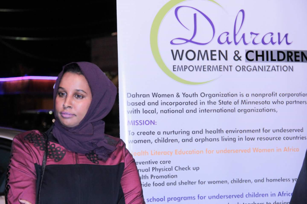 Eine Frau in einem weißen Hemd und blauen Jeans steht entschlossen vor einem Banner mit der Aufschrift "Dahran Frauen & Kinder-Befähigung Organisation", mit einem Auto im Hintergrund.