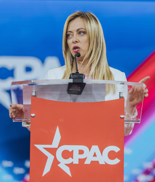 Eine Frau in einem weißen Kleid steht an einem Podium mit einem Mikrofon, unscharfer Hintergrund legt den Fokus auf sie, wahrscheinlich eine Politikwissenschaftlerin.