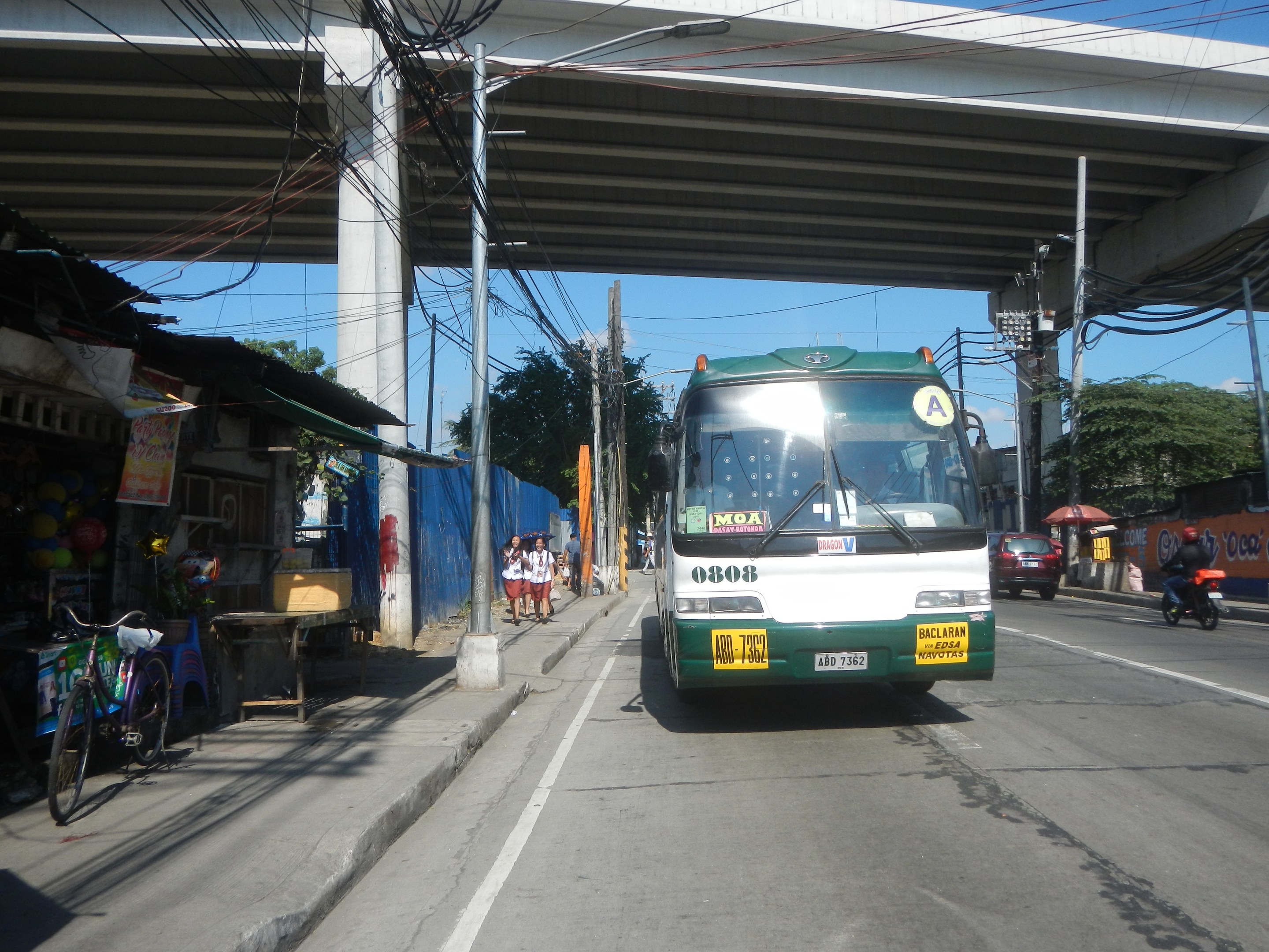 Ein grüner und weißer Bus fährt eine Straße mit Strommasten und Kabeln entlang, vorbei an Geschäften, Ständen und parkenden Fahrrädern, mit Fußgängern auf dem Gehweg und einer Brücke im Hintergrund bei klarem blauem Himmel.