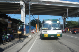 Ein grüner und weißer Bus fährt eine Straße mit Strommasten und Kabeln entlang, vorbei an Geschäften, Ständen und parkenden Fahrrädern, mit Fußgängern auf dem Gehweg und einer Brücke im Hintergrund bei klarem blauem Himmel.