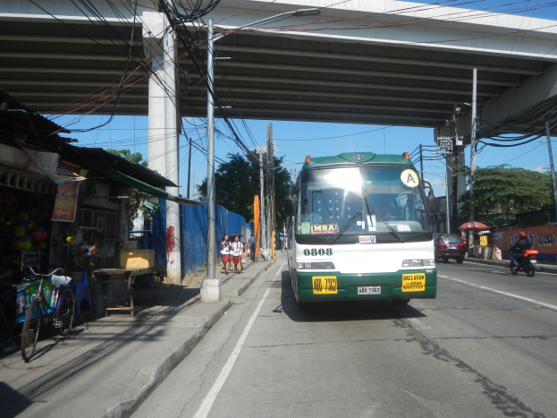 Ein grüner und weißer Bus fährt eine Straße mit Strommasten und Kabeln entlang, vorbei an Geschäften, Ständen und parkenden Fahrrädern, mit Fußgängern auf dem Gehweg und einer Brücke im Hintergrund bei klarem blauem Himmel.