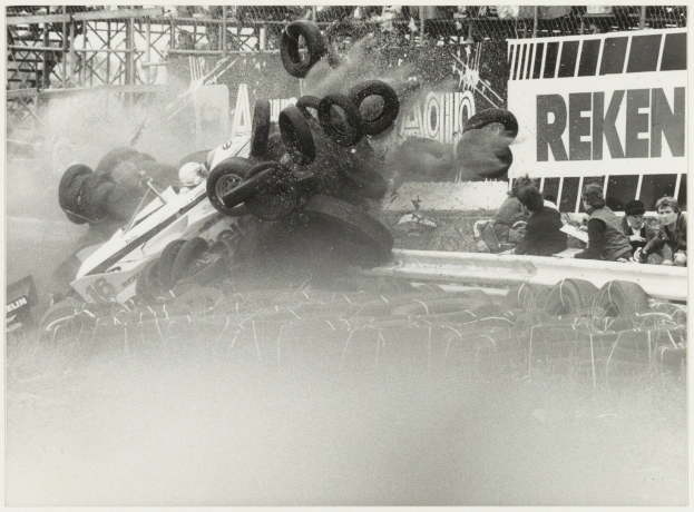 Schwarz-weißes Foto eines verunglückten Rennwagens umgeben von Reifen, mit einer Gruppe von Menschen auf der rechten Seite und einer Tafel mit Text im Hintergrund.