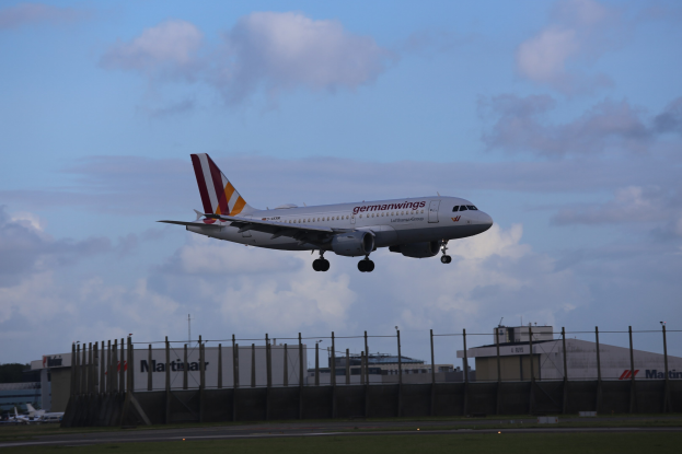 Germanwings Airbus A320-200 startet von Frankfurt Airport mit Gras darunter, einem Zaun im Vordergrund, Gebäuden und Bäumen im Hintergrund und einer bewölkten Himmel