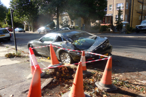 Ein verunglücktes Auto am Straßenrand, umgeben von orangefarbenen Verkehrskegeln, mit Bäumen, Gebäuden und einem klaren blauen Himmel im Hintergrund.