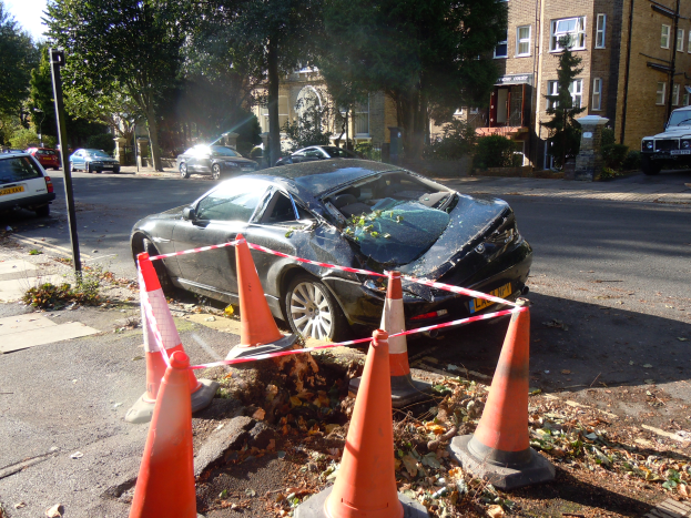 Ein verunglücktes Auto am Straßenrand, umgeben von orangefarbenen Verkehrskegeln, mit Bäumen, Gebäuden und einem klaren blauen Himmel im Hintergrund.
