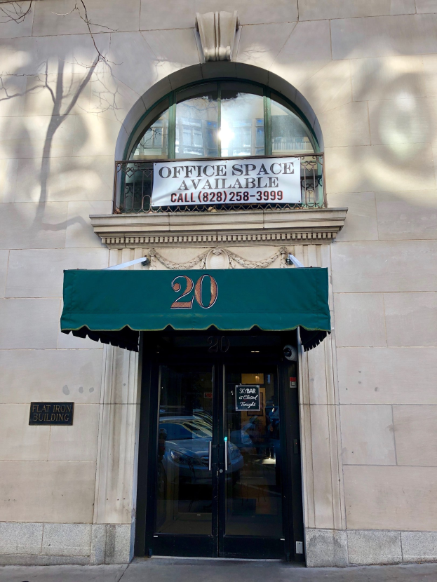 Ein Gebäude mit einer Glastür, einem grünen Vordach, einem Schild mit der Aufschrift "Büroräume zu vermieten", einem Geländer und einem Baum links.