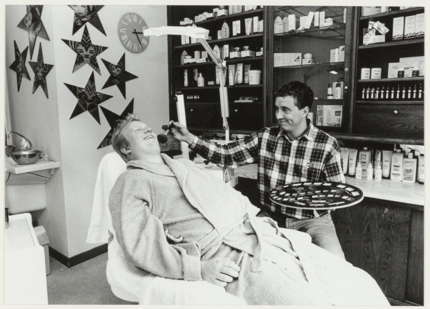 Ein Mann lächelt in einem Zahnarztstuhl, während ein anderer Mann neben ihm steht und einen Teller hält, mit einem Waschbecken, einer Uhr an der Wand und Regalen mit Flaschen im Hintergrund.