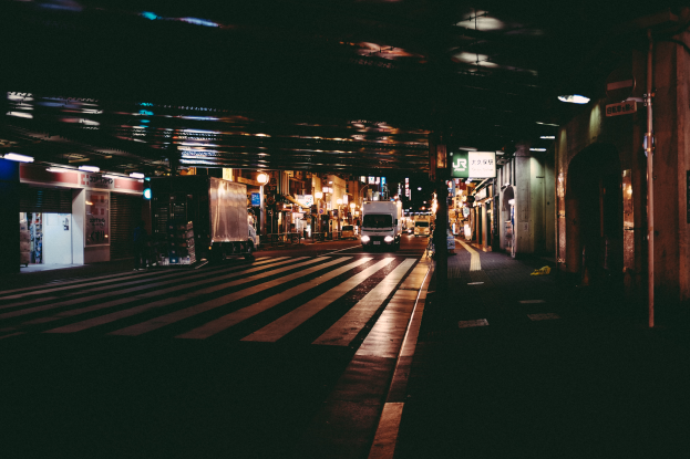 Eine Stadtstraße bei Nacht mit Fahrzeugen auf der Straße, Gebäuden auf beiden Seiten, beleuchteter Fläche und einer Brücke im Hintergrund.