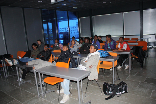 Eine Gruppe von Menschen, die auf Stühlen in einem Klassenzimmer sitzen und auf einen Schreibtisch blicken, mit Fenstern und Vorhängen an der Rückwand.