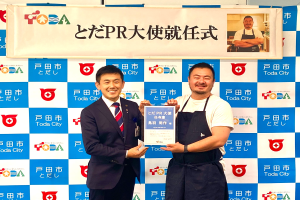 Zwei lachende Männer mit einem Zertifikat, im Hintergrund ein Banner mit der Aufschrift "Toda City Wins PR Award" und einem Foto einer Person.