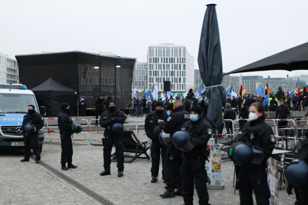 Eine Gruppe von Polizeibeamten steht vor einer Menge von Demonstranten in Berlin, Deutschland, mit Barrikaden, Zelten und Fahnen im Vordergrund und Gebäuden im Hintergrund und einem klaren Himmel.