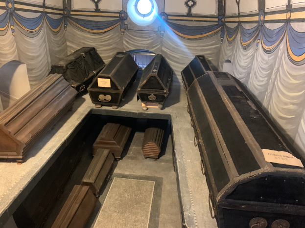 Ein Raum im Holocaust-Museum in Berlin, Deutschland, gefüllt mit Holzsärgen auf Tischen, mit einer Vorhangwand und einer Lampe im Hintergrund.