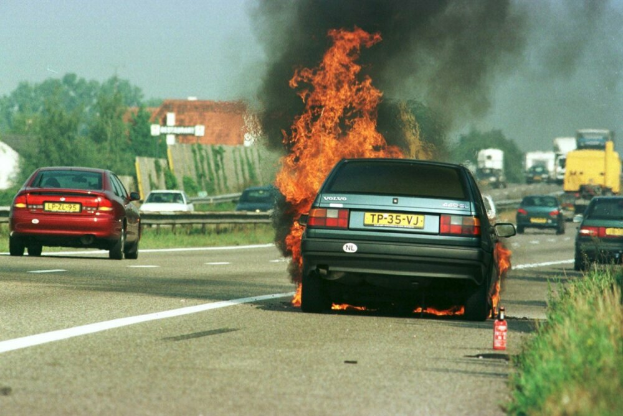 Ein Auto steht in Flammen am Straßenrand, umgeben von anderen Fahrzeugen, mit Bäumen, Gebäuden und einem klaren blauen Himmel im Hintergrund und einem Feuerlöscher auf der rechten Seite im Gras.