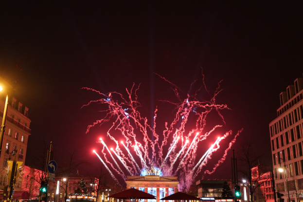 Eine belebte Stadtstraße während einer Silvesterfeier in Berlin, voller Menschen, Fahrzeuge und festlicher Beleuchtung von Gebäuden und Feuerwerk am Himmel.
