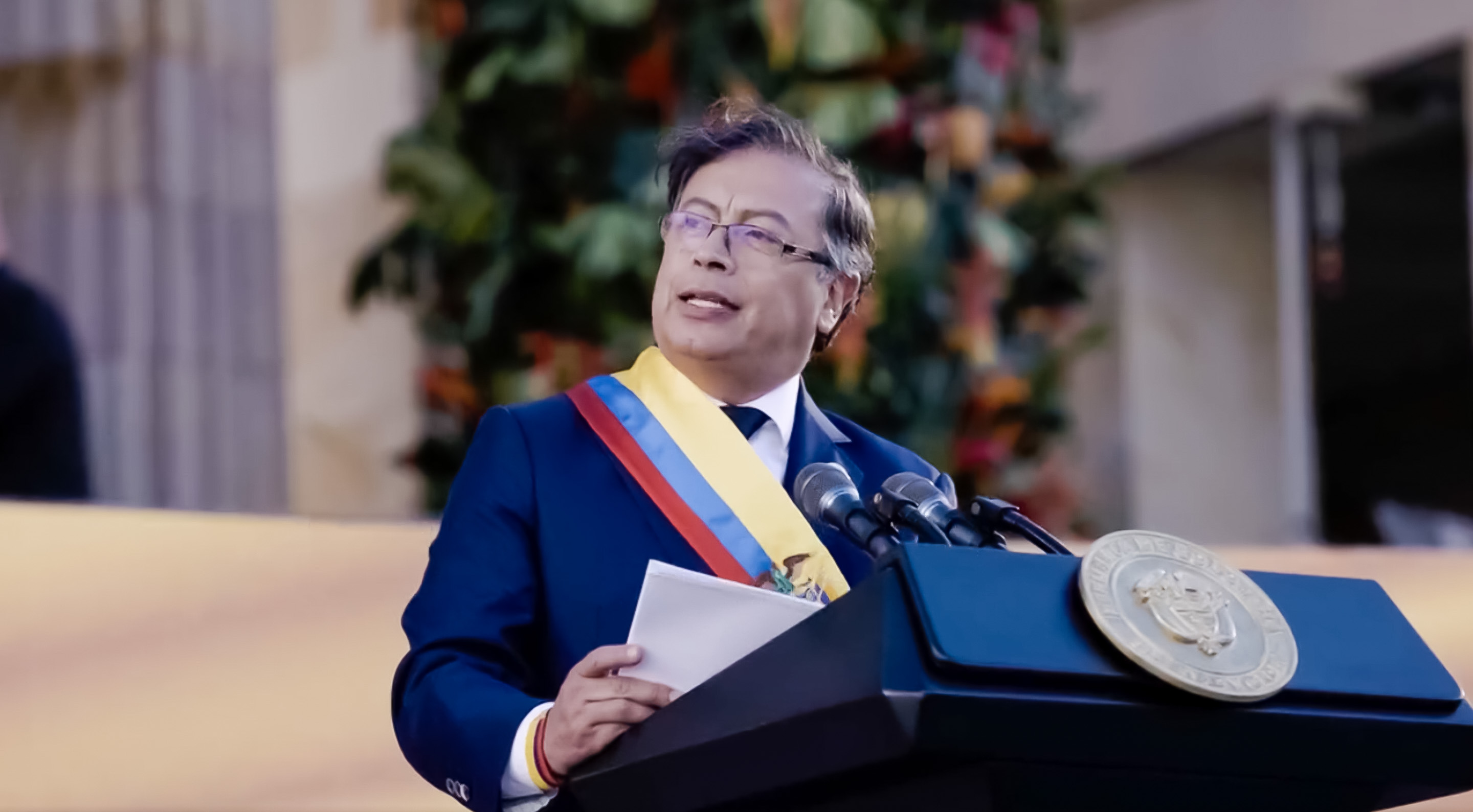 Ein Mann in einem blauen Anzug und Krawatte steht an einem Rednerpult, hält Papiere und spricht in ein Mikrofon, mit einer Pflanze und einer Wand dahinter, was auf eine formelle Umgebung hindeutet.